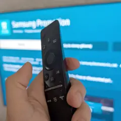 Dłoń trzymająca pilot Samsunga na tle niebieskiego ekranu telewizora z widocznymi ikonami aplikacji.