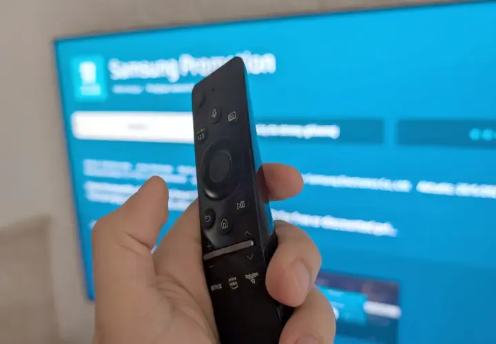Dłoń trzymająca pilot Samsunga na tle niebieskiego ekranu telewizora z widocznymi ikonami aplikacji.