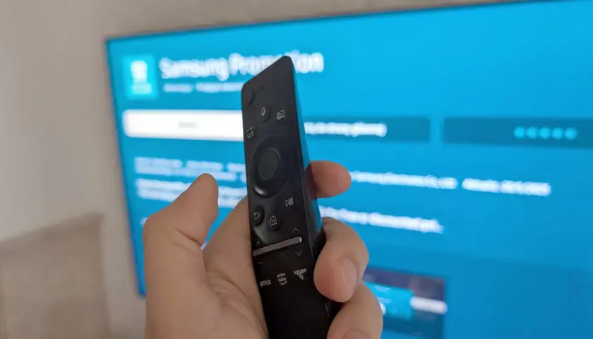 Dłoń trzymająca pilot Samsunga na tle niebieskiego ekranu telewizora z widocznymi ikonami aplikacji.
